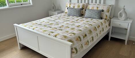 A white bed with a leaf-patterned bedspread, two matching pillows, a nightstand with a vase, and a window with a view of greenery.