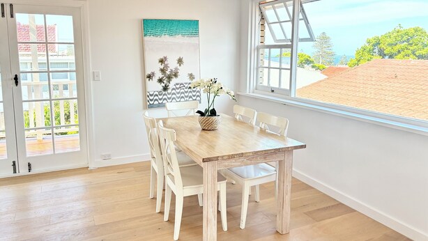 A dining area with a wooden table and white chairs, a painting on the wall, and a view of a beach through a large window. - *New Collaroy 3 B/R Northern Beaches Spacious & Renovated Beach Getaway Collaroy (Collaroy)