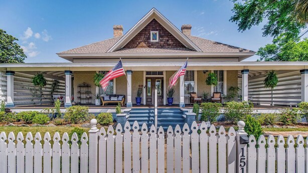 A two-story house with a covered porch, American flags, and a white picket fence. - Pensacola • 1BR • Sleeps 2 • Close to Downtown (Pensacola)