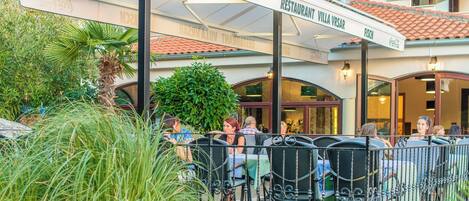 An outdoor seating area with tables and chairs, a Coca-Cola branded awning, and a sign for a restaurant named 'Villa Vrsar Grill'.