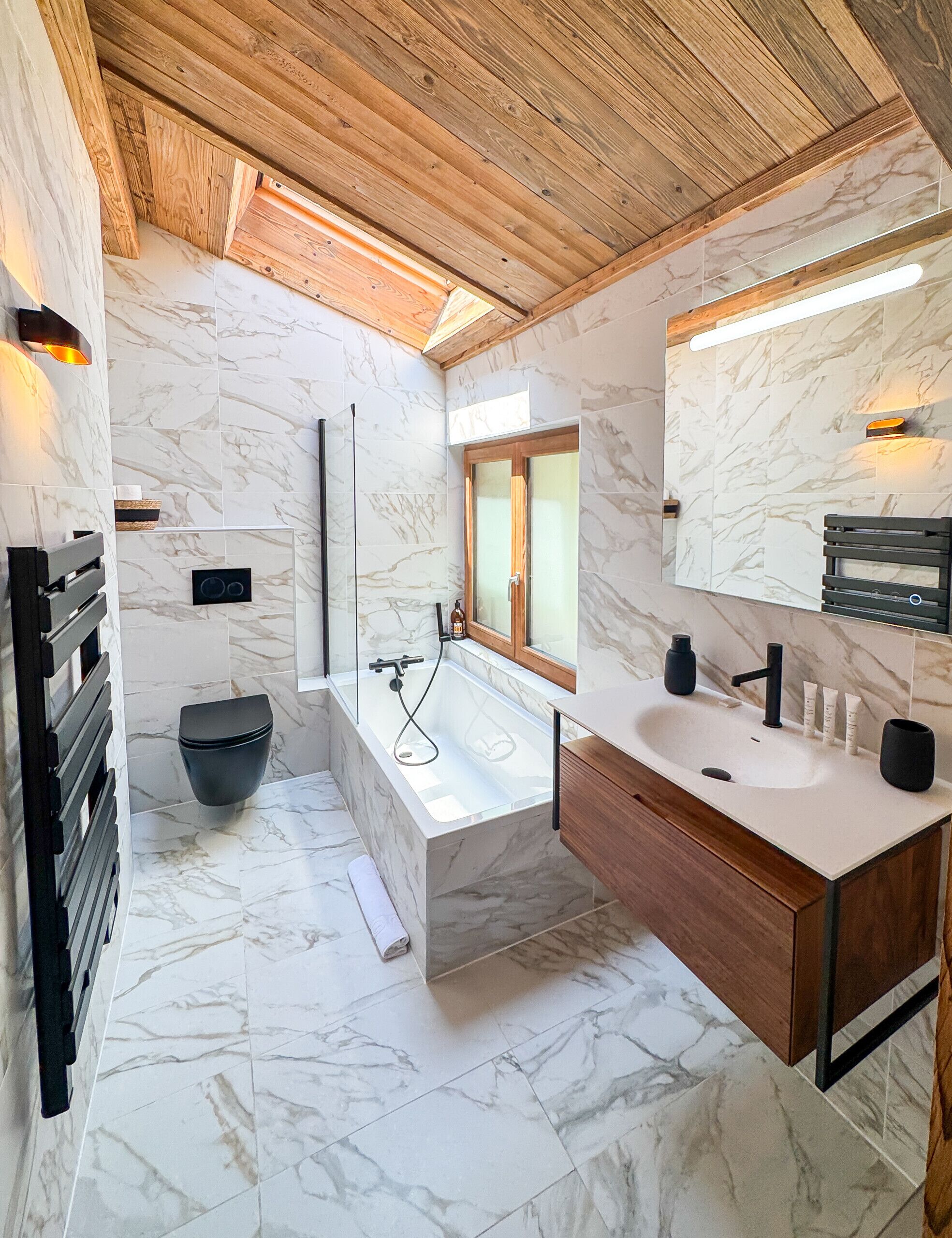 Une salle de bain moderne avec un sol en marbre, une baignoire, un lavabo avec un miroir et des poutres en bois au plafond.