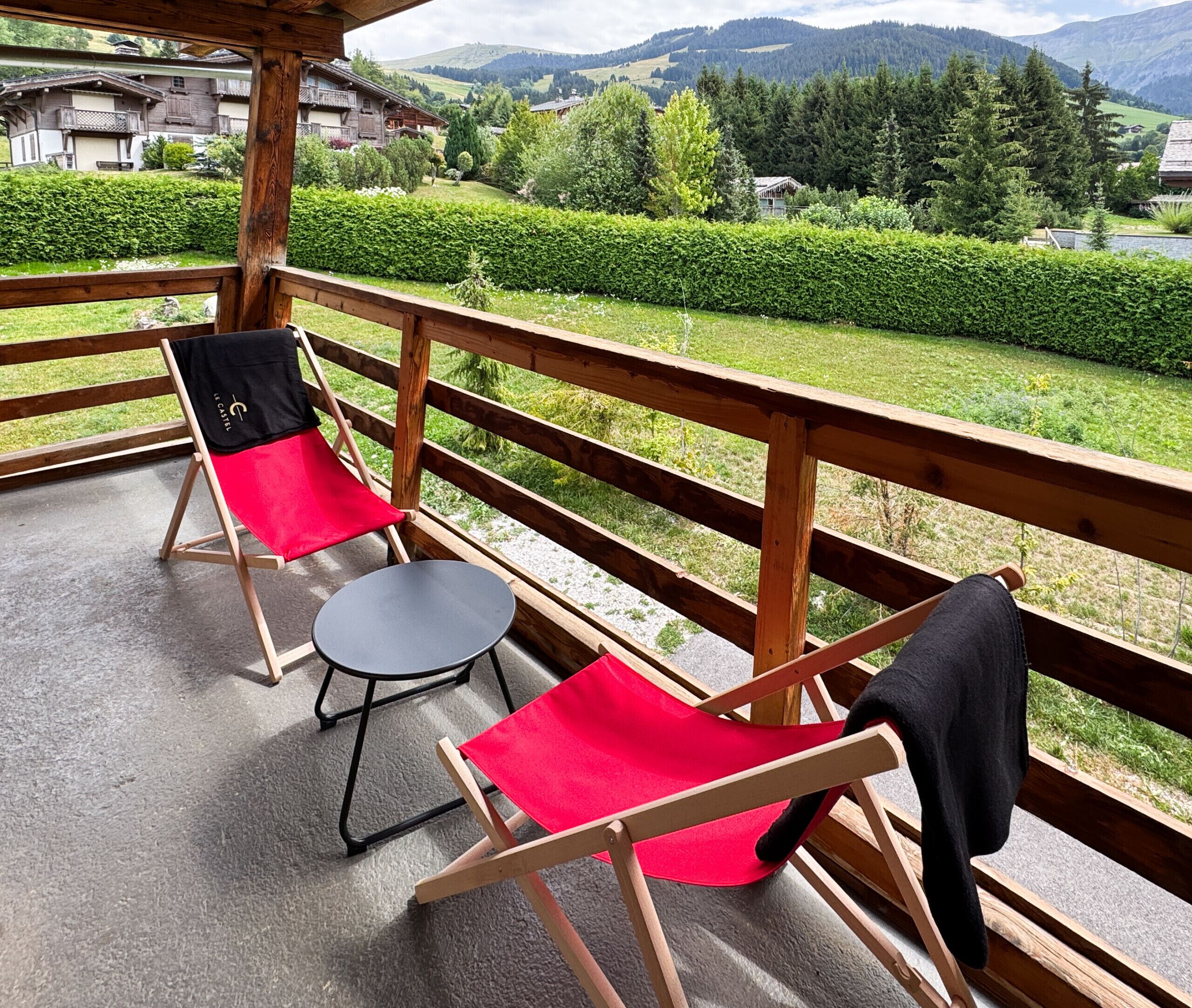 Un balcon avec deux chaises et une petite table, donnant sur un paysage verdoyant avec des bâtiments en arrière-plan.