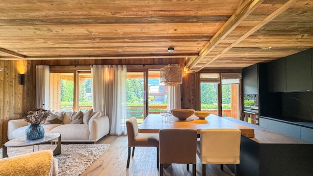 Ein modernes Interieur mit Holzbalkendecke, einer Essecke mit Tisch und Stühlen, einer Couch und einem großen Fenster mit Blick auf Grünflächen.