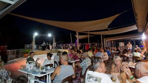 Un área de comedor al aire libre con mesas y sillas, un escenario con artistas y un toldo sobre la zona.