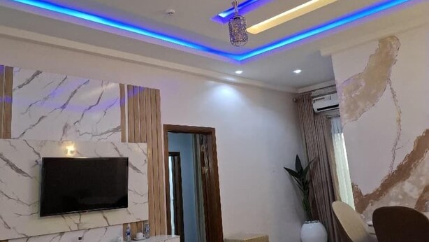 A modern living room with a flat-screen TV mounted on a marble wall, a ceiling with blue LED lighting, and a round coffee table. - Nice-mason Serene and Suites (Benin City)