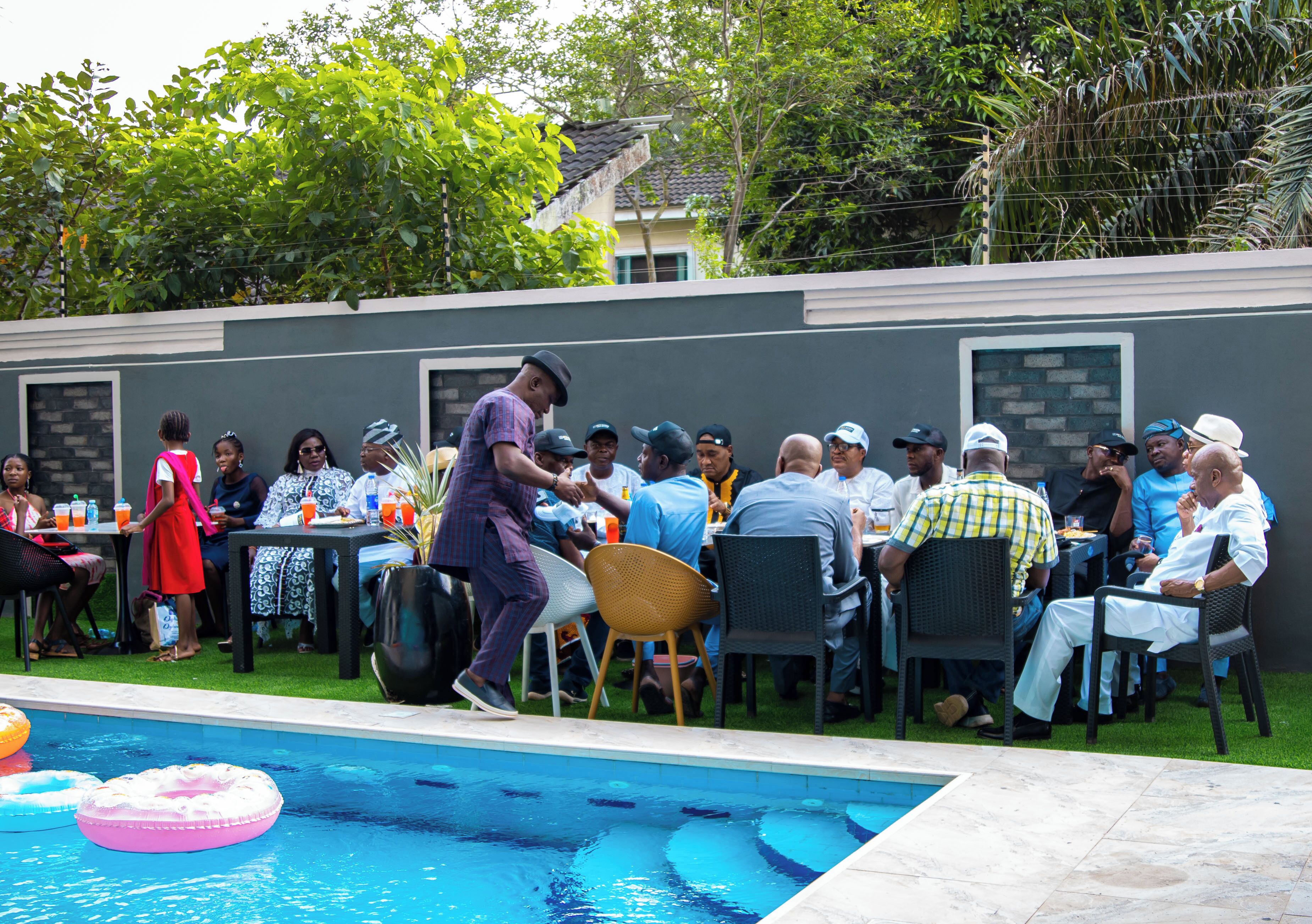A poolside gathering with people seated at tables, a pool with inflatable rings, and a building with a tiled roof in the background.