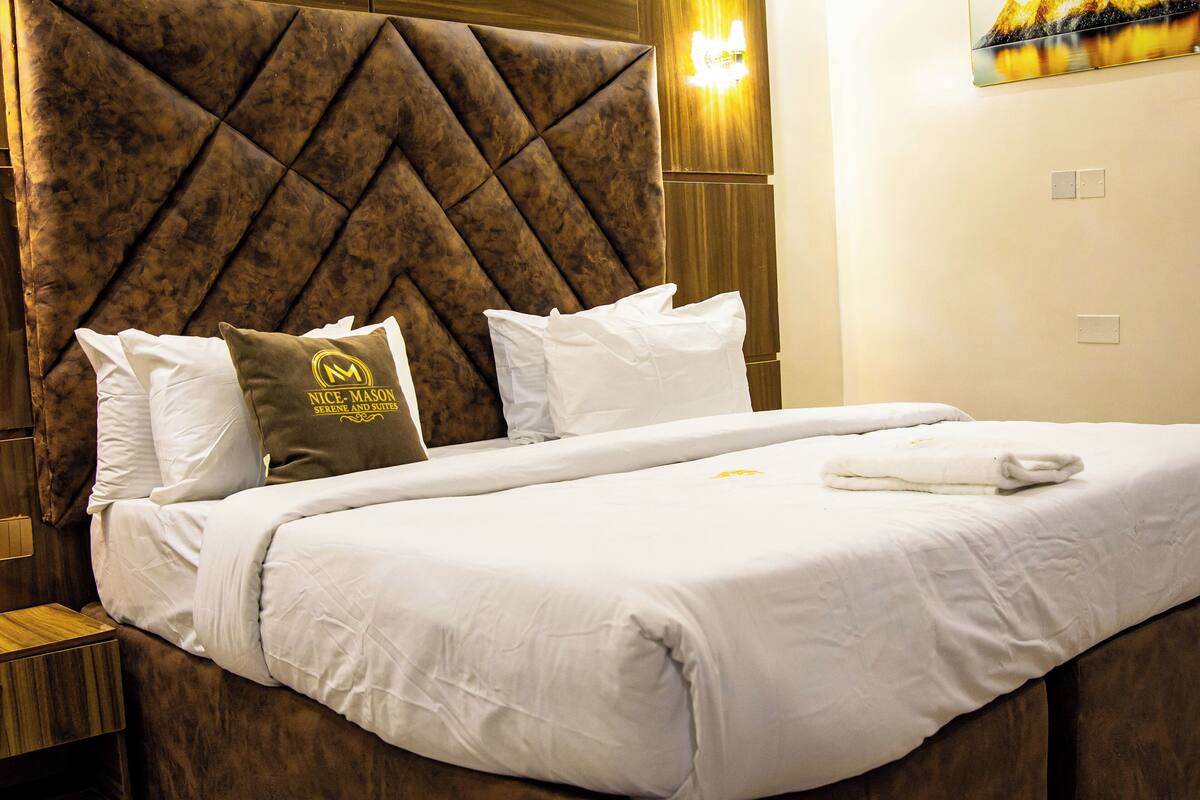 Un lit bien fait, avec du linge de lit blanc et une tête de lit marron. On peut voir des oreillers sur le lit, dont l’un porte un logo.