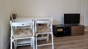 Una habitación con una mesa blanca y dos sillas blancas con cojines beige, un mueble de entretenimiento negro con televisor, y suelo de madera.