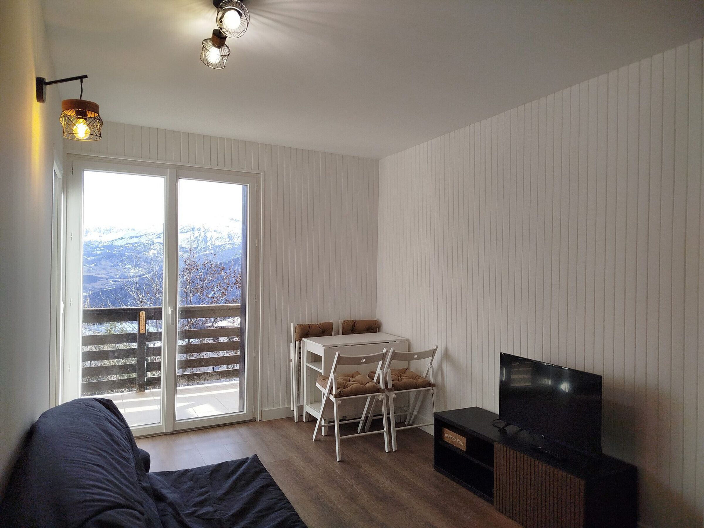 Ein modernes Wohnzimmer mit einer Couch, einem Fernseher, einem Esstisch mit Stühlen und einem Balkon mit Blick auf schneebedeckte Berge.