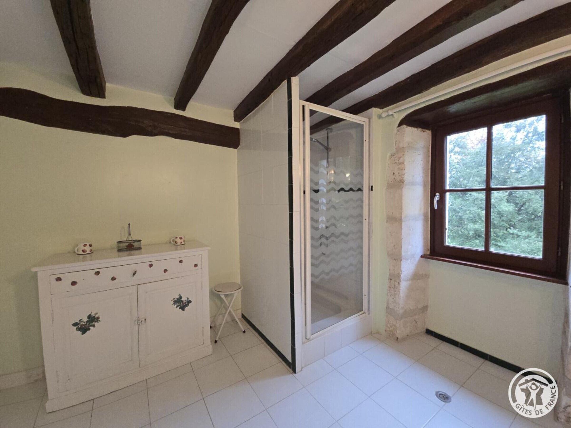 Une pièce avec un meuble blanc, un petit tabouret, une porte-fenêtre coulissante et une fenêtre donnant sur des arbres.