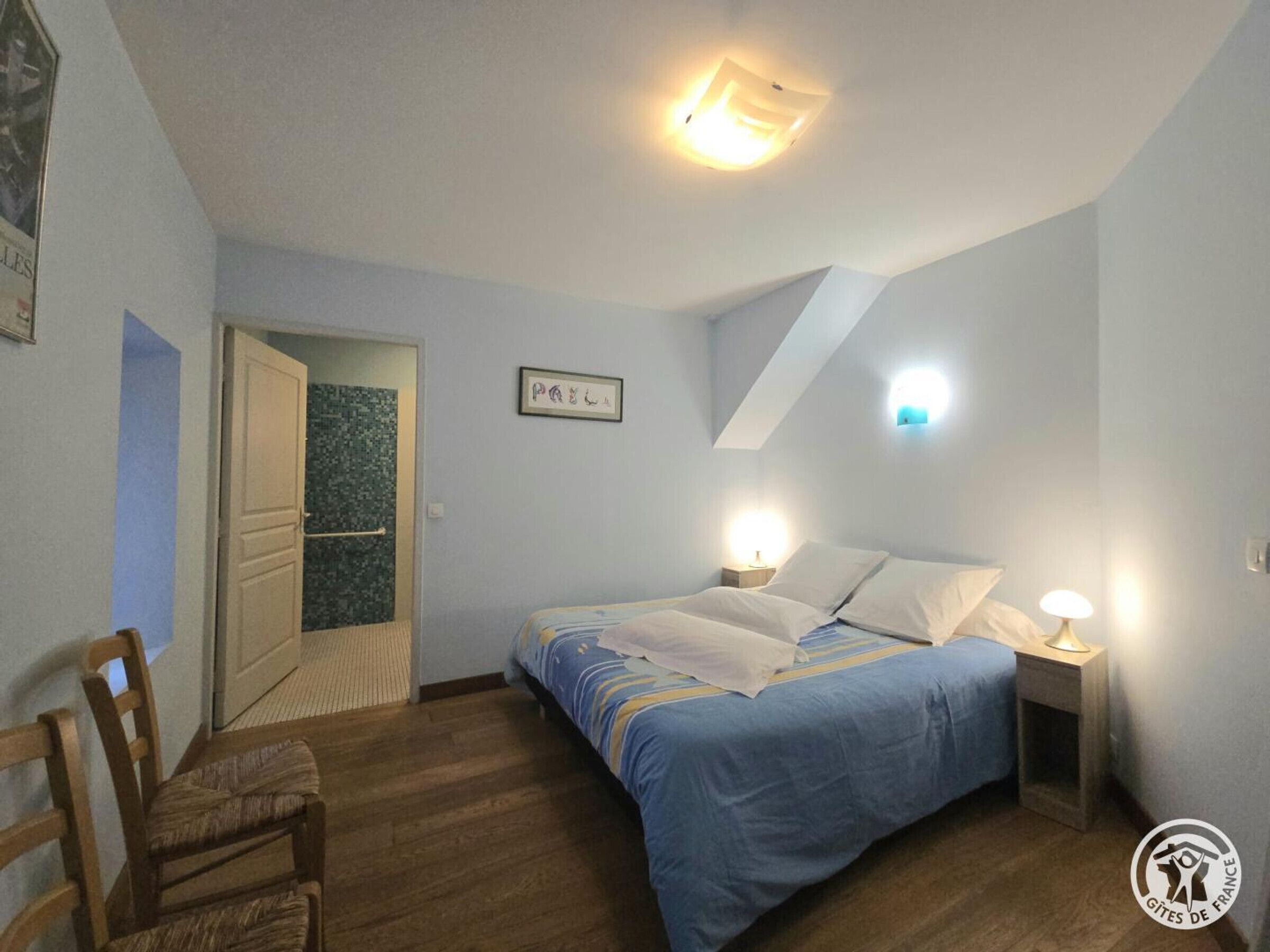 Een slaapkamer met een bed, bedlampen, een houten stoel en een deur naar een andere kamer.