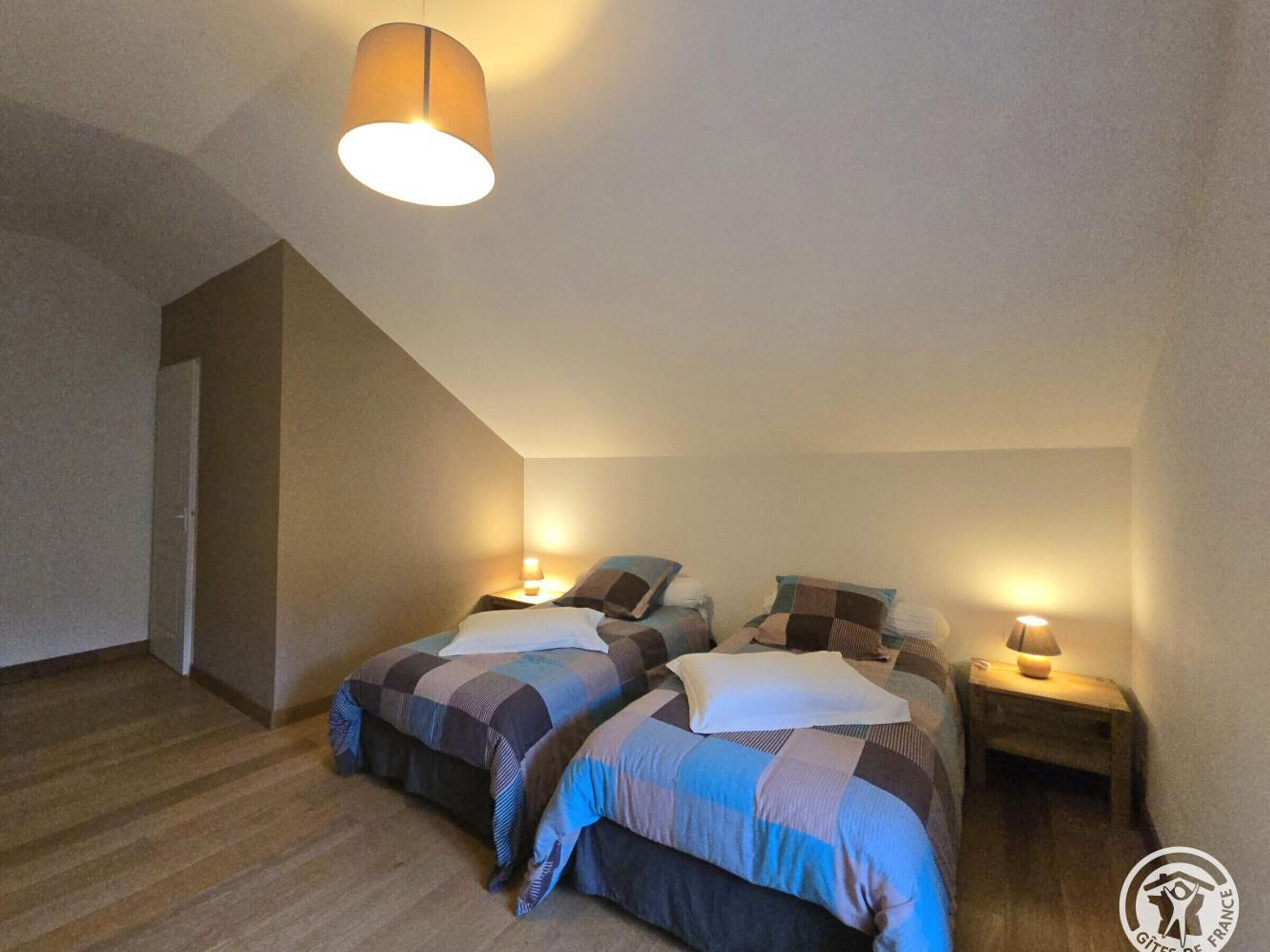 Une chambre avec deux lits, chacun recouvert d’une couette et de coussins à carreaux, un luminaire suspendu et une petite table de chevet avec une lampe.