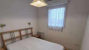 A bedroom with a wooden bed, a bedside table, a wall-mounted heater, and a window with sheer curtains.
