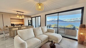 Un soggiorno moderno con un divano ampio, una zona pranzo e una cucina. Una grande finestra offre vista sul mare e sulle montagne.