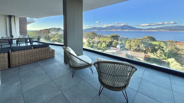 Un balcone moderno con zona pranzo e due poltrone in vimini, con vista su un paesaggio costiero che include montagne e un tratto d'acqua.