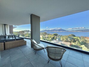 Un balcone moderno con zona pranzo e due poltrone in vimini, con vista su un paesaggio costiero che include montagne e un tratto d'acqua.