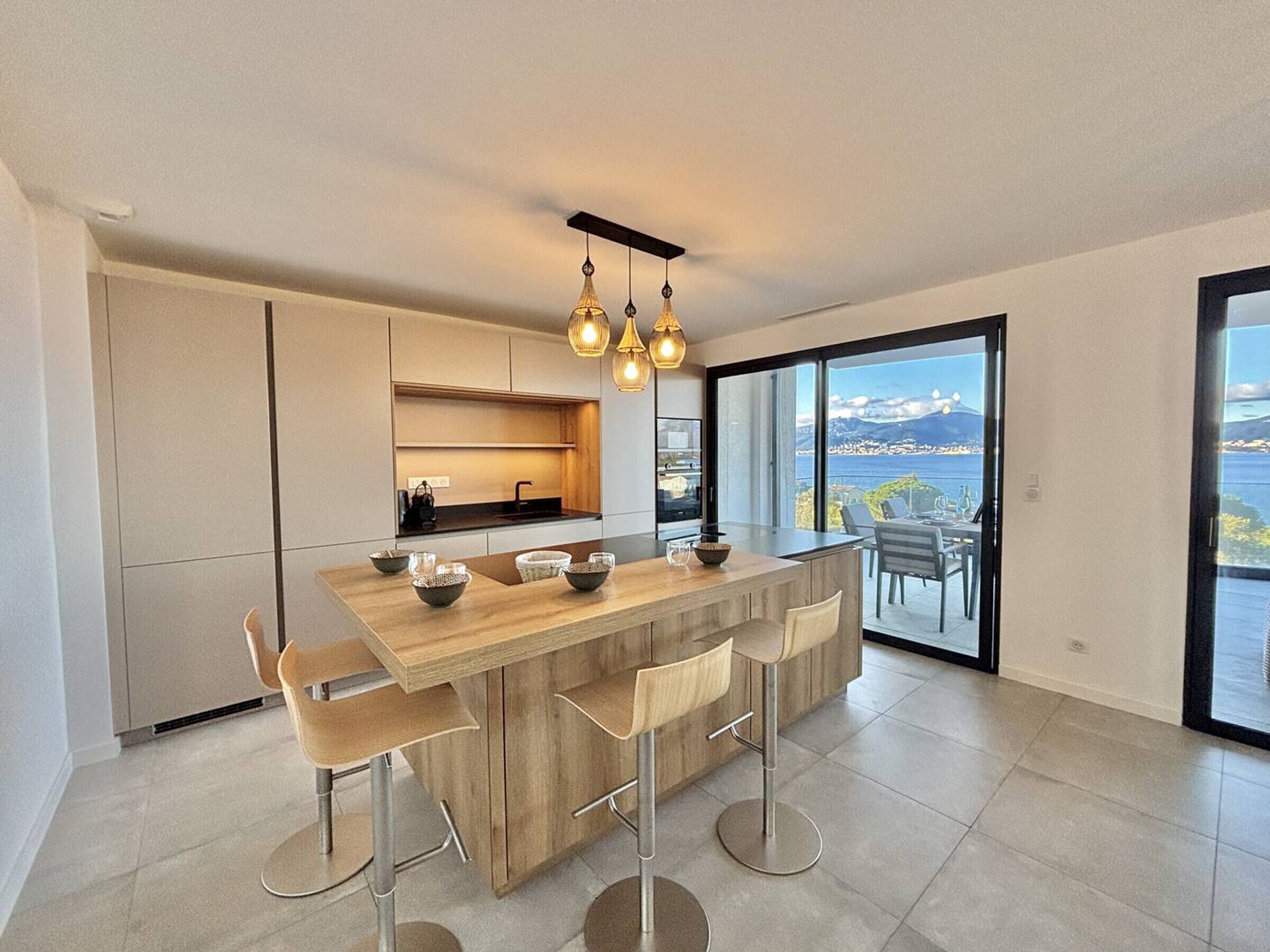 A modern kitchen with a central island, bar stools, and a view of the sea through large windows.