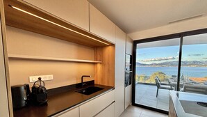 Una cucina moderna con un piano di lavoro nero, un lavello integrato e una mensola in legno. Offre una vista panoramica su un corpo d'acqua e un paesaggio lontano.