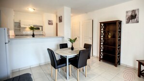 Cocina moderna con armarios blancos, un microondas y una planta sobre la encimera. Hay una zona de comedor con una mesa y sillas, y una estantería de madera contra la pared.