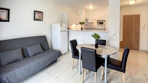 Una sala moderna con un sofá gris, una mesa de comedor con cuatro sillas, y una zona de cocina con refrigerador, microondas y armarios.