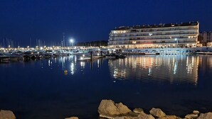 Un port de plaisance de nuit, avec des bateaux amarrés, un grand bâtiment doté de nombreux balcons, et une côte rocheuse au premier plan.