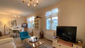 Un soggiorno con televisione, una poltrona blu, un divano bianco, un tavolino da caffè e un lampadario.