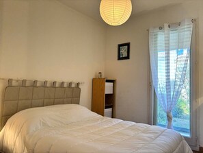 A bedroom with a bed, a nightstand, a wooden cabinet, a ceiling light, and a window with curtains. - Hyères T3 Garden, 4 Pers., Parking, Near Beaches (Hyères)
