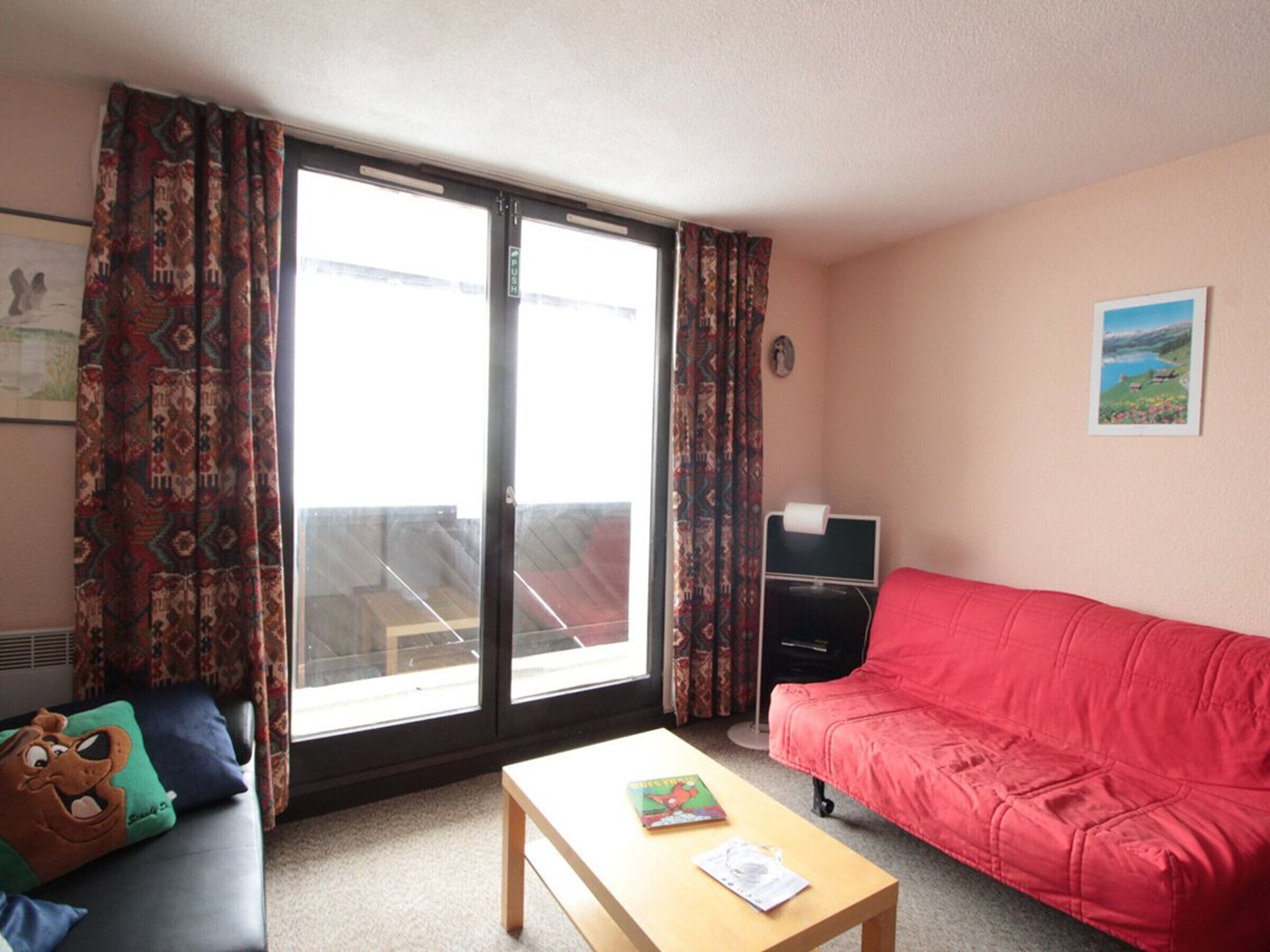 A living room with a red sofa, a wooden coffee table, patterned curtains, and a window with a view.