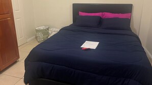 Una cama bien hecha con una colcha azul oscuro y detalles rosados, un cesto de basura pequeño y una puerta de madera.