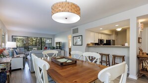 Un coin repas avec une table en bois et des chaises blanches, une cuisine en arrière-plan et une grande fenêtre qui laisse entrer la lumière naturelle.