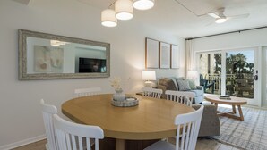 Un coin repas avec une table ronde en bois, des chaises blanches, un lustre, un grand miroir et un téléviseur fixé au mur.