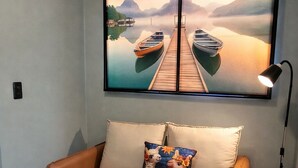 Een kamer met een beige bank, een kleurrijk kussen, een lijst met een foto van boten op een steiger en een lamp.