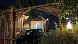 Une entrée fermée par un portail, avec une voiture garée à l’intérieur, un petit bâtiment doté d’un toit en pente et un feuillage dense au premier plan.