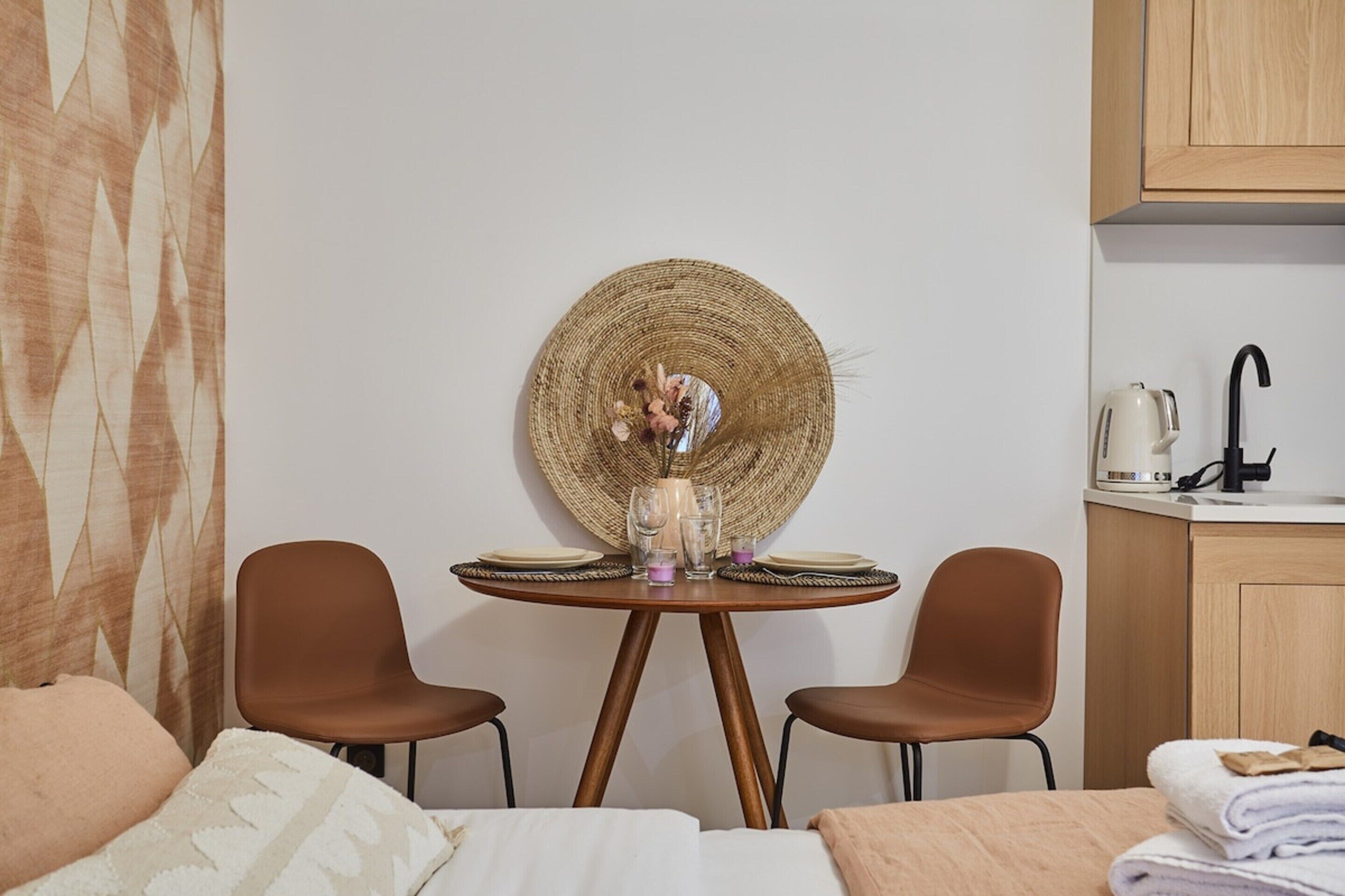 A room with a round table set for two, a wicker wall hanging, and a kitchenette area with a kettle and sink.