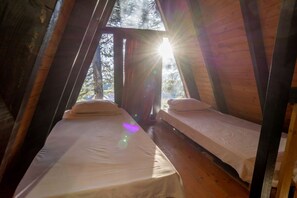 Ein gemütliches Schlafzimmer im Dachgeschoss mit zwei Betten, Holzbalken und Blick auf Bäume durch ein Fenster.