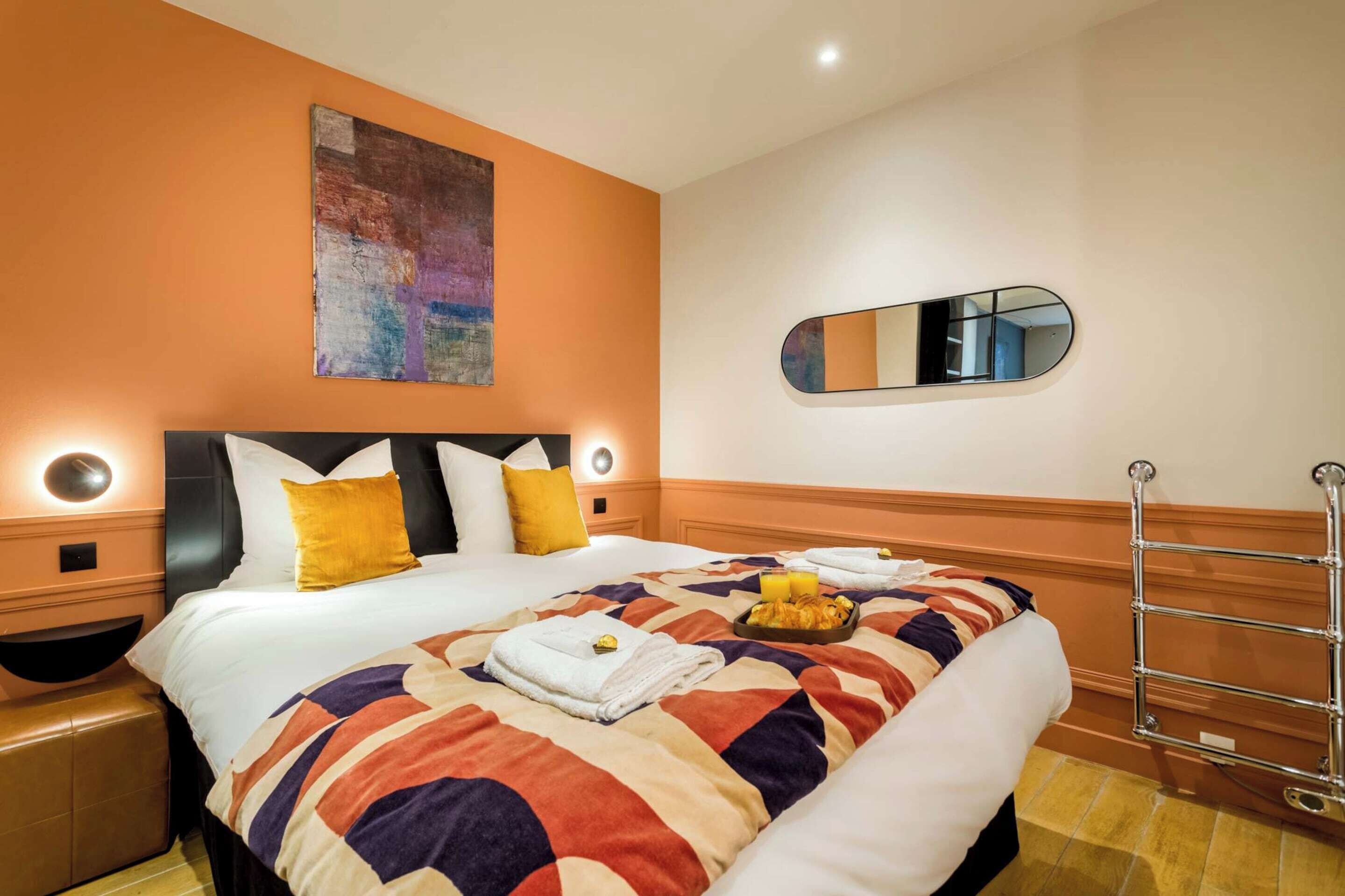 A hotel room with a bed, a painting, a mirror, and a towel rack.