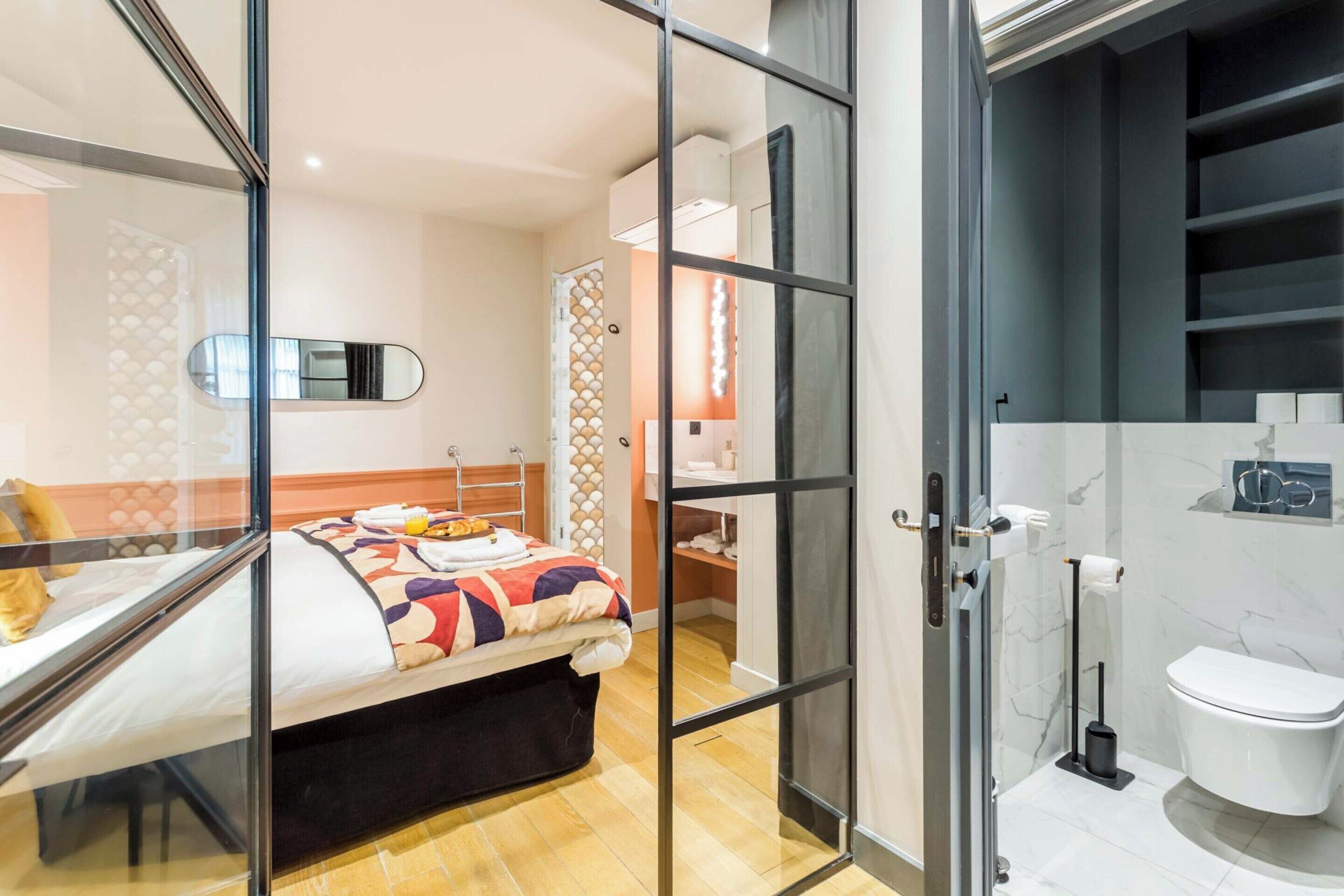 A modern hotel room with a bed, a glass partition, a bathroom with a toilet and sink, and a mirror.
