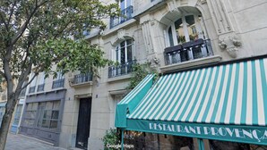 A multi-story building with balconies, a restaurant with a striped awning, and a tree in the foreground. - Place des Vosges, Apartment With Air Conditioning (Paris)