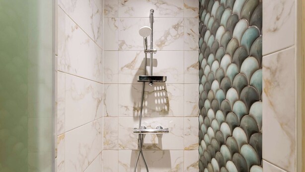 A shower with a handheld showerhead and a wall-mounted faucet, set against a tiled wall with a decorative tile pattern. - Place des Vosges, Apartment With Air Conditioning (Paris)