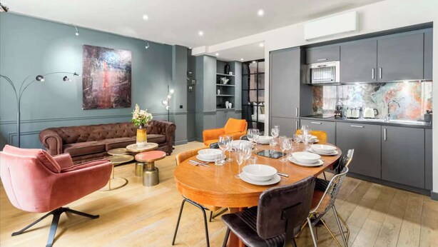 A modern living room with a sofa, a dining table set for four, and a kitchen with grey cabinets and a built-in oven. - Place des Vosges, Apartment With Air Conditioning (Paris)