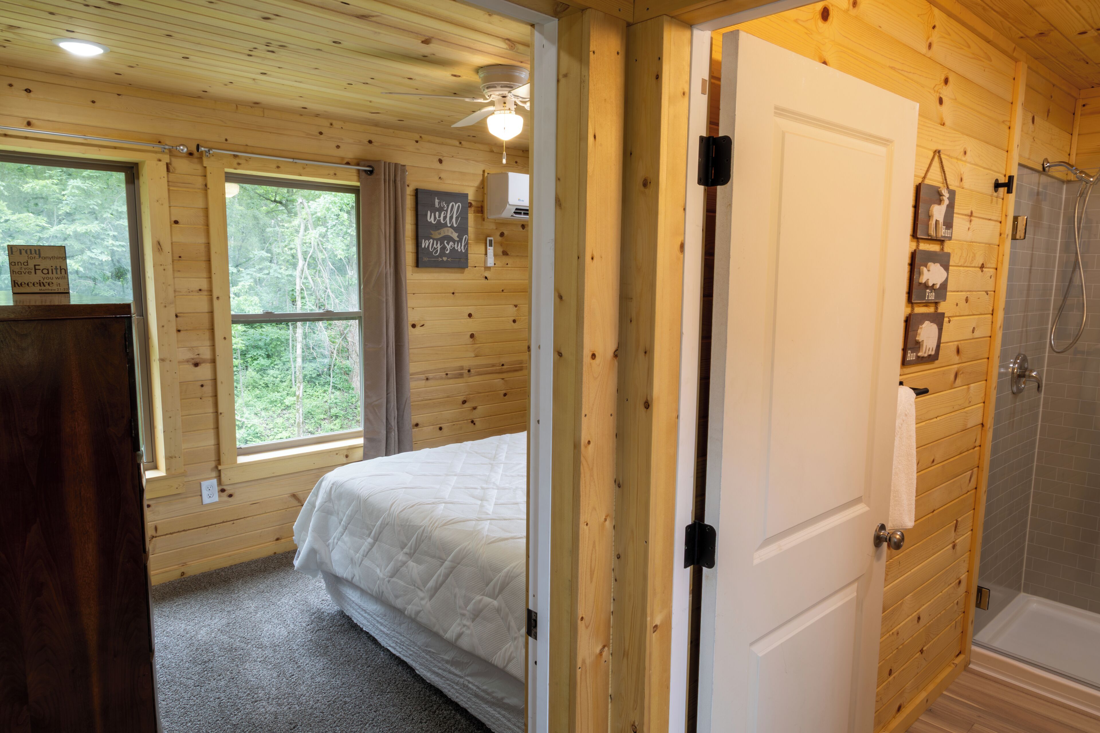 Un dormitorio de cabaña de madera con una cama, un ventilador de techo, una ventana y una puerta que da a un baño con ducha.