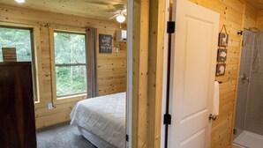 Quarto de cabana de madeira com cama, ventilador de teto, janela e porta que dá para um banheiro com chuveiro.