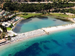 Vista aérea de uma área costeira com praia, piscina e resort com vários edifícios e guarda-sóis.