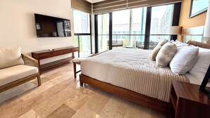 Un dormitorio con una cama grande, un escritorio, una silla, un ventilador de techo y ventanas amplias con vistas a edificios.