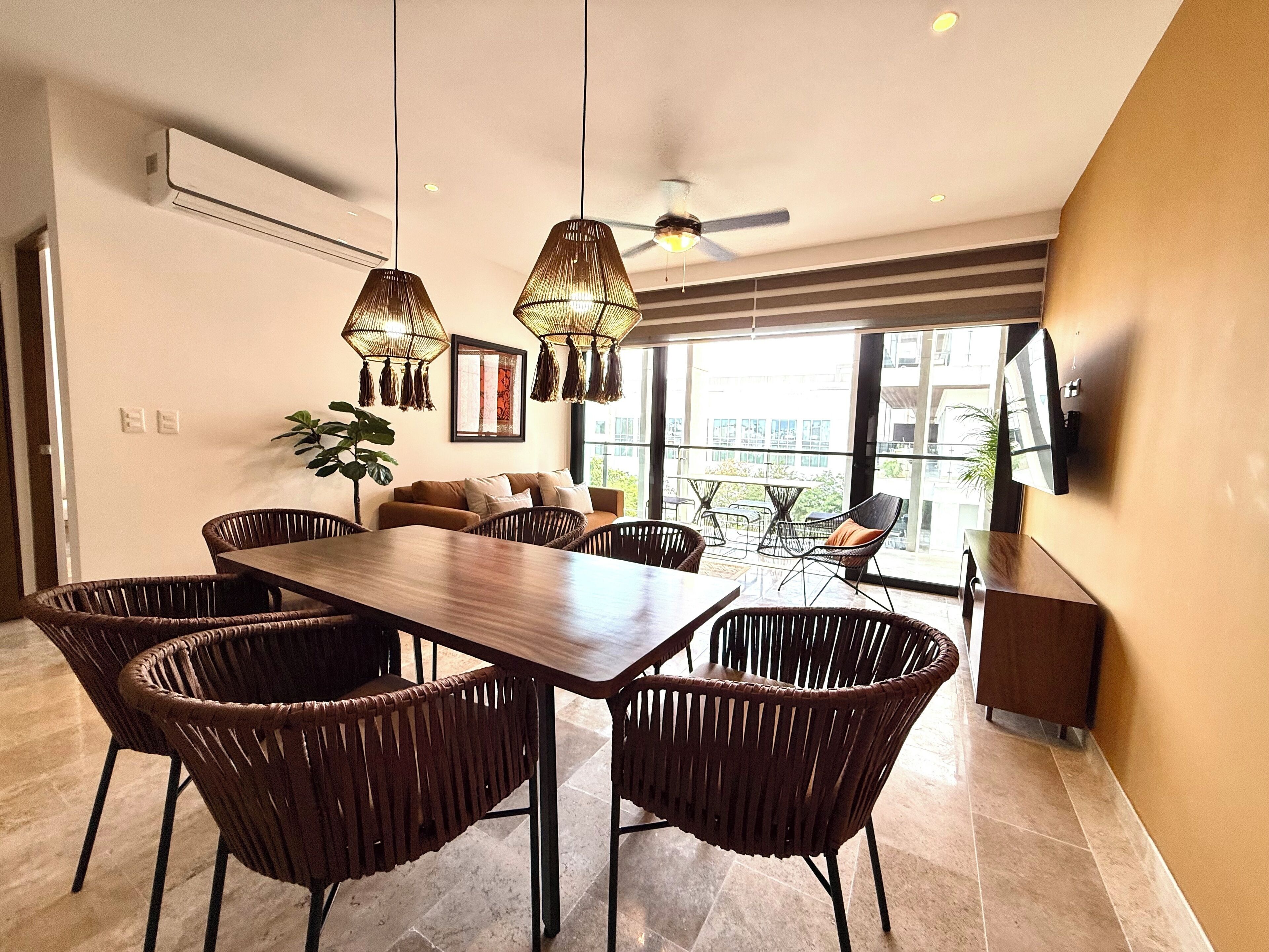 Un espace repas moderne avec une table en bois et des chaises en osier, un ventilateur de plafond et une grande fenêtre donnant sur la ville.