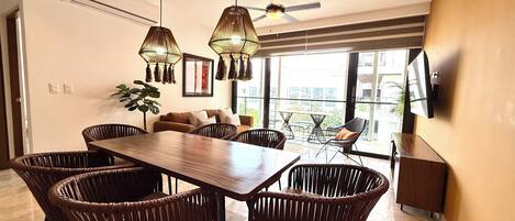 Un espace repas moderne avec une table en bois et des chaises en osier, un ventilateur de plafond et une grande fenêtre donnant sur la ville.