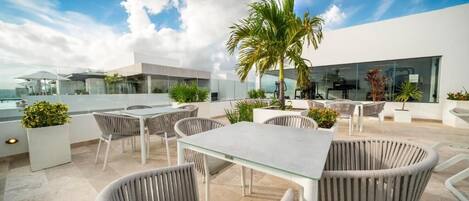 Un patio al aire libre con mobiliario moderno, que incluye sillas de mimbre y una mesa de vidrio, rodeado de plantas en macetas y una palmera.