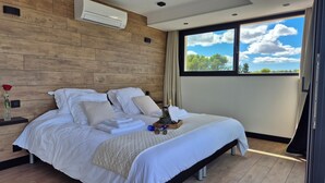 A modern bedroom with a wooden headboard, a large bed with white linens, a bedside table with a vase of flowers, and a window with a view of the sky and trees. - L'Eden Bleu in Langlade, upscale romantic suite with private SPA. (Langlade)