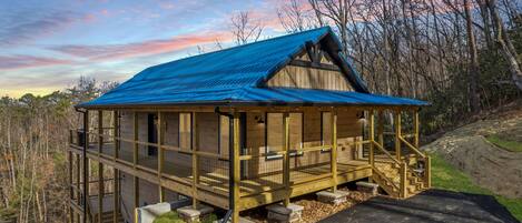 A wooden cabin with a blue roof, a balcony, and a staircase leading to the entrance.
