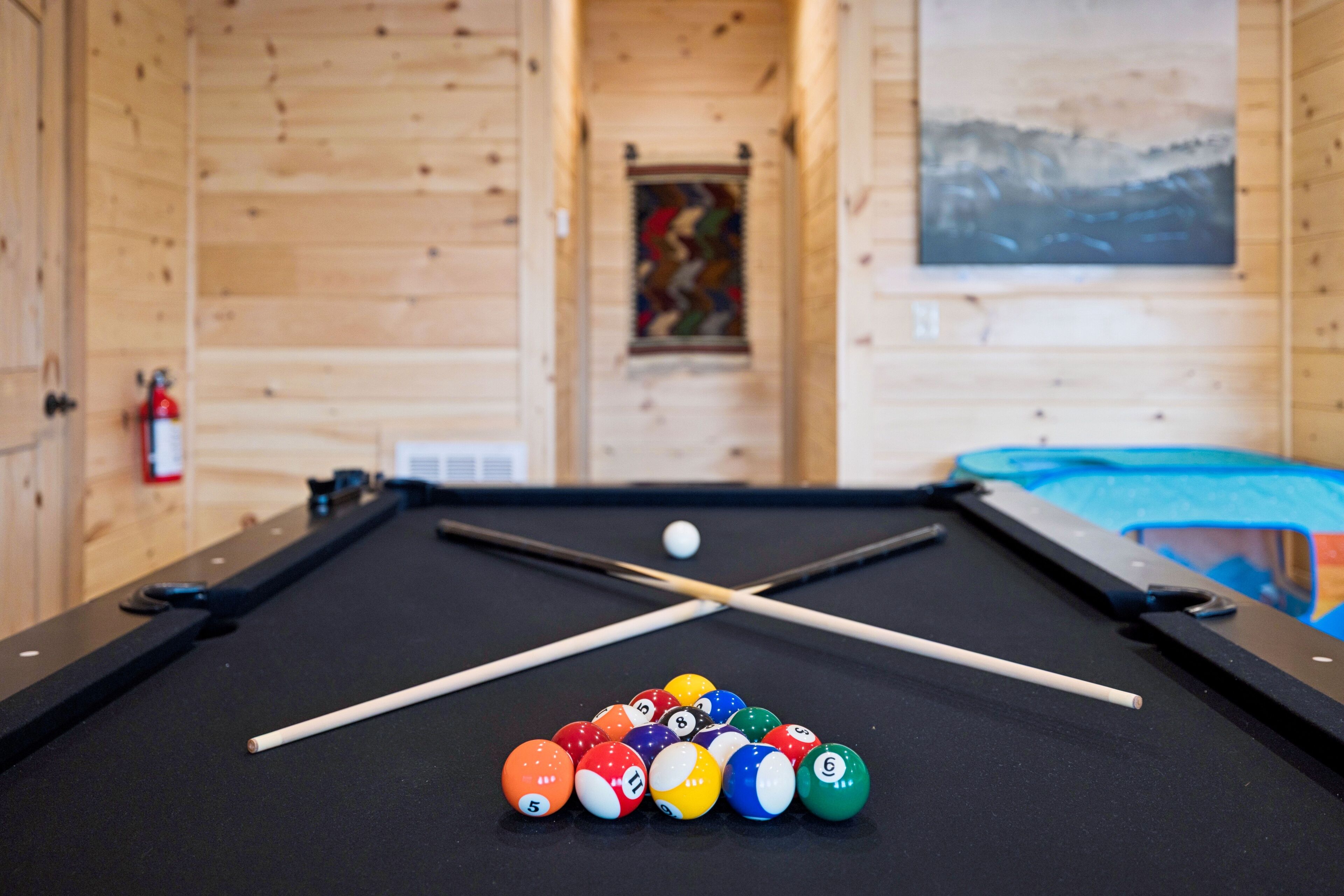 A pool table with billiard balls and cues, wooden walls, and a fire extinguisher.